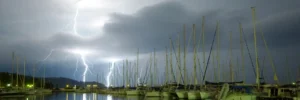 Mehrere Blitzeinschläge hinter einem Hafen mit vertäuten Segelbooten während eines nächtlichen Unwetters, mit Spiegelungen im ruhigen Wasser und dramatischen Wolken am Himmel
