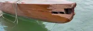 Vorderer Bereich einer klassischen Yacht aus Holz mit schweren strukturellen Schäden am Bug, gesplittertem Holz und herausgebrochenen Planken, schwimmend auf ruhigem, grünlichem Wasser