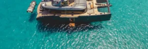 Katamaran-Yacht mit Beibooten und Sonnenliegen liegt in türkisblauem Wasser vor Anker, von oben fotografiert.